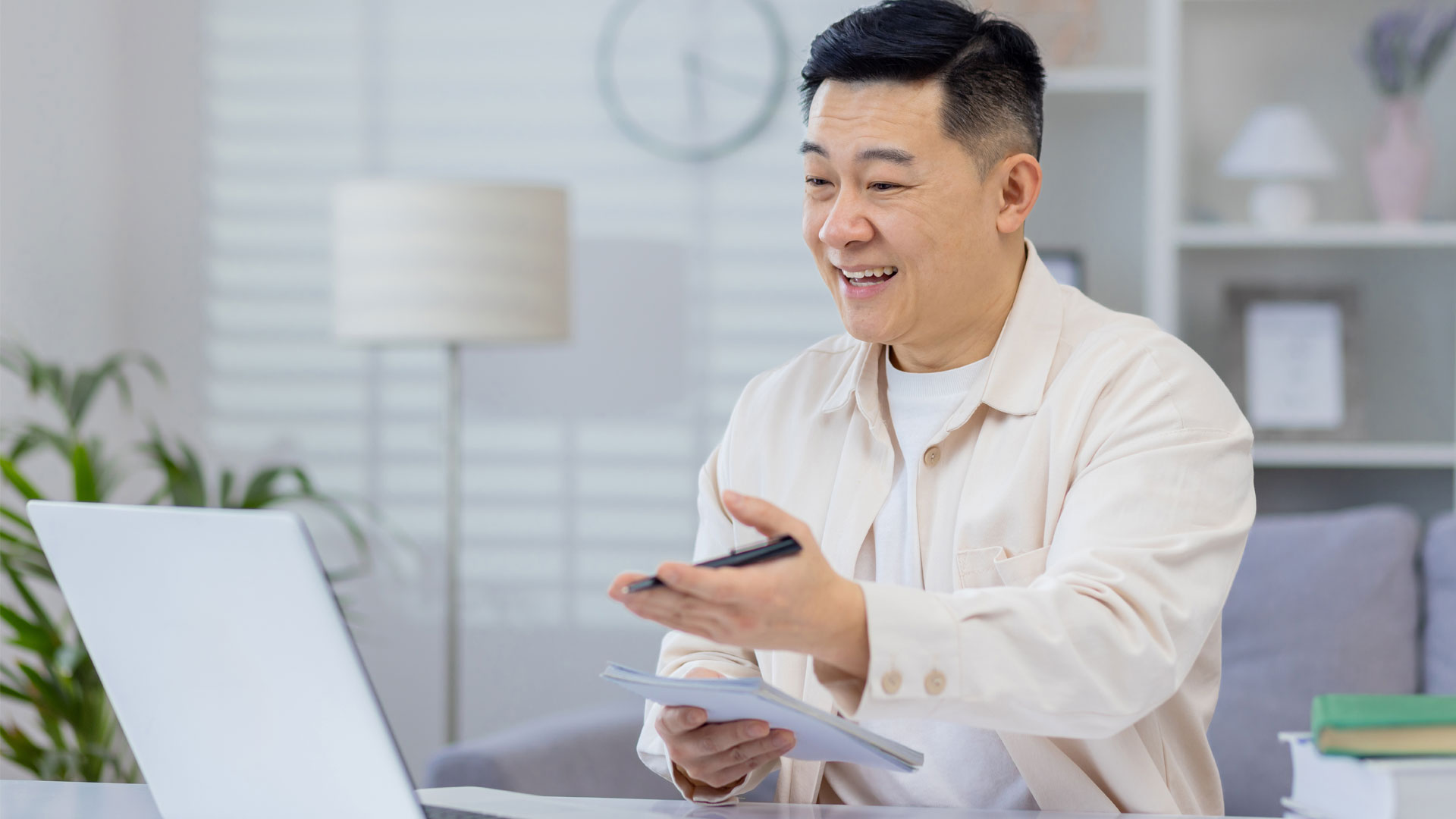 Asian male talking while holding pen and paper looking at a laptop