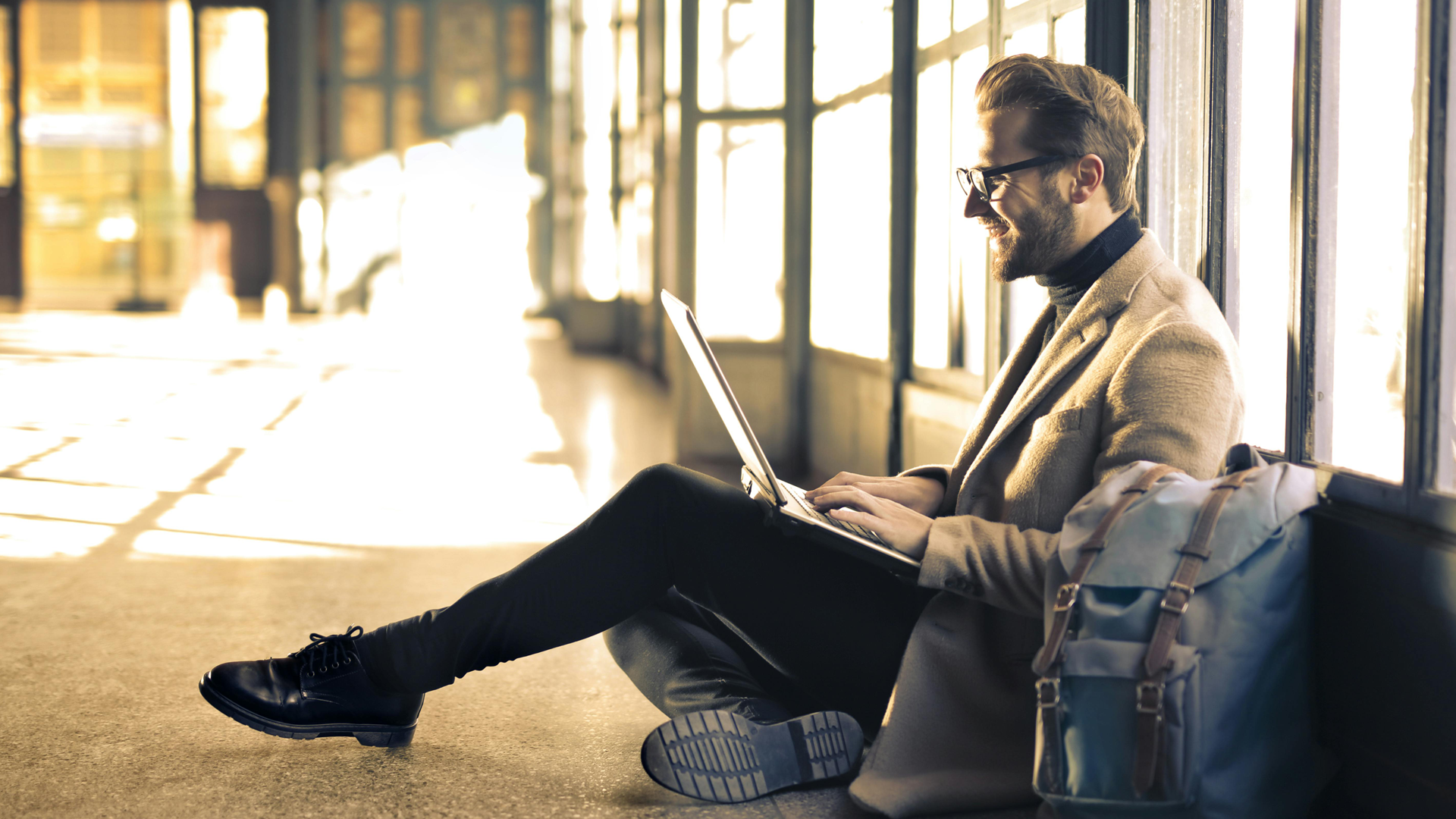 A man person sitting on the floor inside a building by windows looking at his laptop